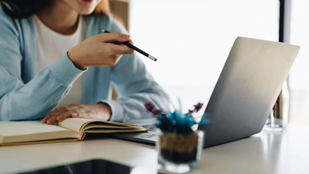 Marketing für Autor:innen Autorin arbeitet mit aufgeschlagenem Buch und Laptop am Schreibtisch