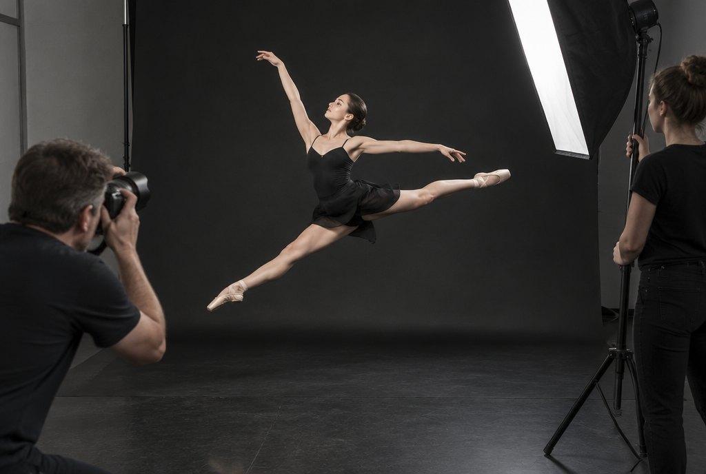 Ein Fotograf fotografiert eine Tänzerin im Sprung vor dunklem Hintergrund im Studio, eine Assistentin hält das Licht (KI-generiert)